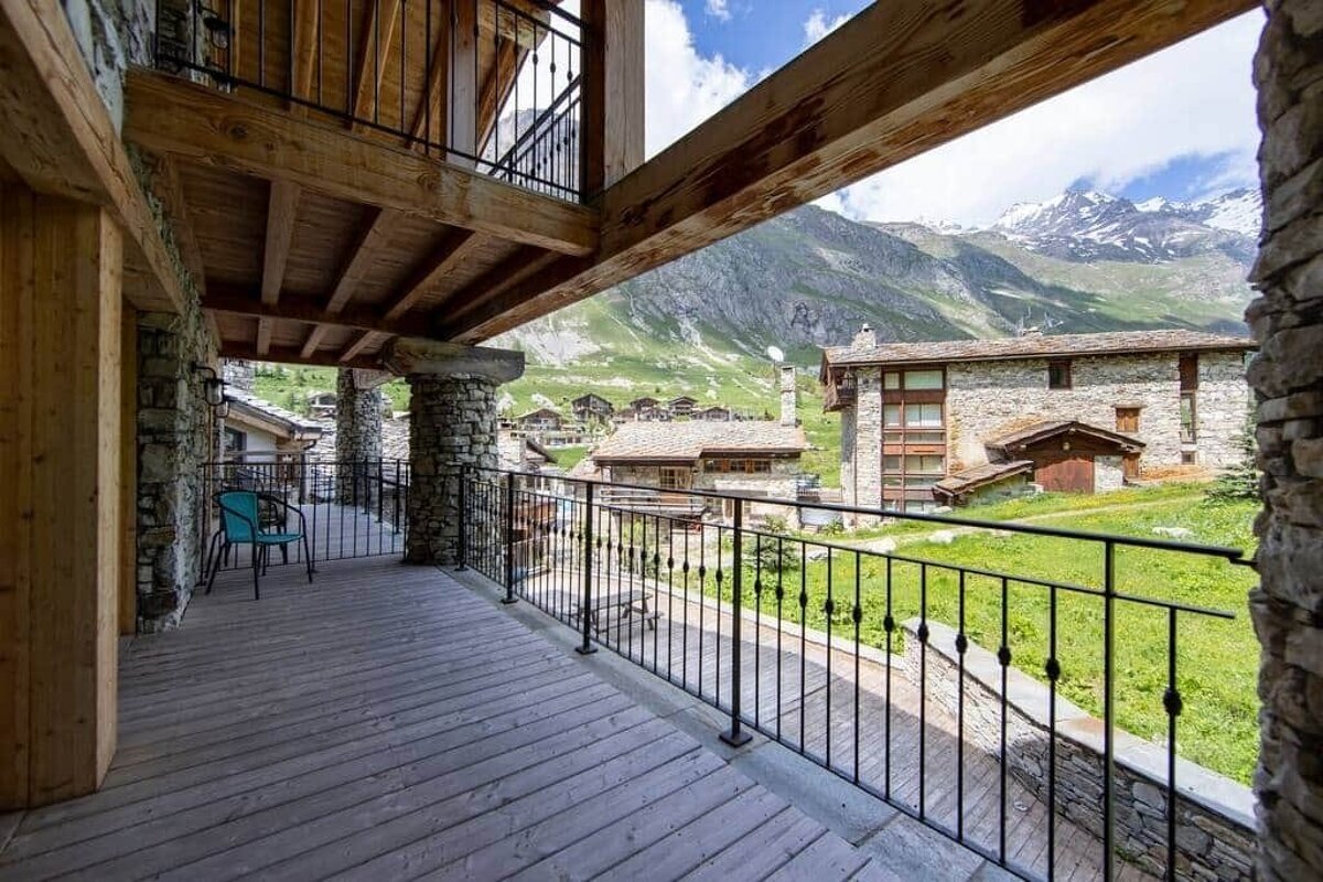 A rustic wooden balcony with stone pillars offers a view of a mountain village, green valley, and snowy peaks under a blue sky.