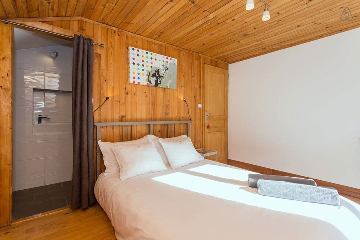 A cozy, rustic-style bedroom features wood-paneled walls and ceiling, a white bed, and a shower visible behind a curtain. Modern art hangs above the bed.