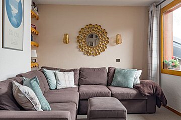 A cozy living room features a large L-shaped taupe sofa with light blue pillows. A golden sunburst mirror and sconces adorn the wall above it.