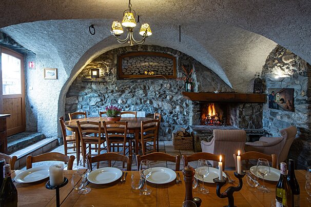 A warm, rustic dining room features stone walls, an arched ceiling, a crackling fireplace, and two wooden tables set for a cozy meal.