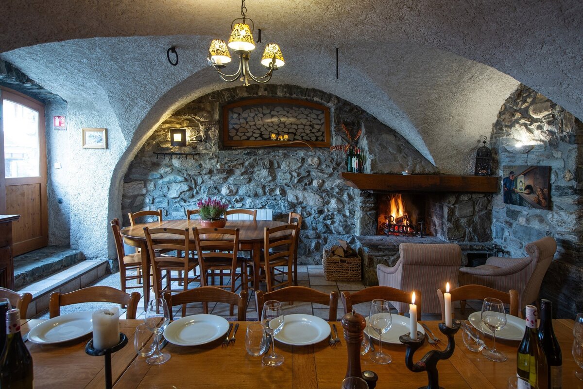 A warm, rustic dining room features stone walls, an arched ceiling, a crackling fireplace, and two wooden tables set for a cozy meal.