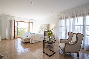 A bright, elegant bedroom with a large bed, balcony to green views, cozy seating, white decor, and tiled floors.