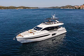 A white luxury yacht floats on calm blue water with a scenic, hilly coastline under a clear sky.