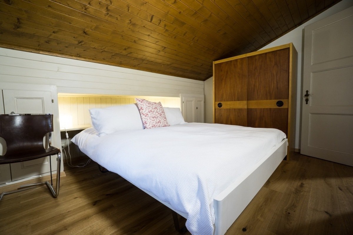 A bedroom with a white bed, sloping wooden ceiling, wooden floor, and a large wooden wardrobe.
