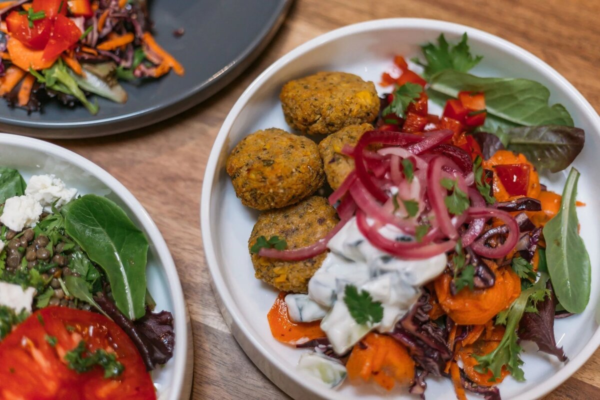 A vibrant bowl of golden falafel, pickled red onions, creamy sauce, and colorful mixed greens. Other healthy salads are in the background.
