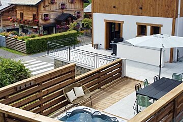 An elevated outdoor terrace with a hot tub, dining area, and lounge chair. Beyond, a charming wooden chalet, green hedges, and a street are visible.