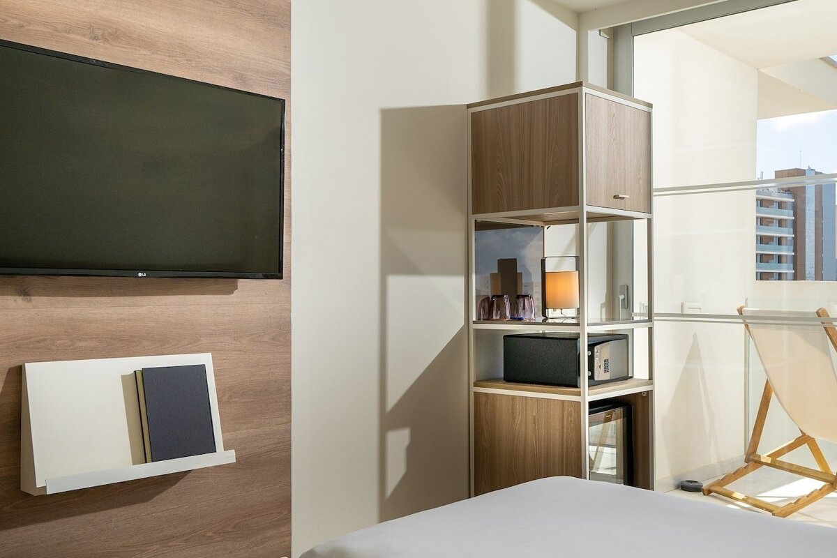 A cozy modern room with a wall-mounted TV, a shelving unit containing a safe and mini-fridge, and a bed. A window reveals an urban view.
