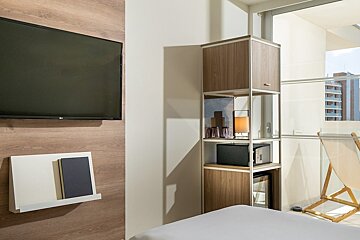 A cozy modern room with a wall-mounted TV, a shelving unit containing a safe and mini-fridge, and a bed. A window reveals an urban view.