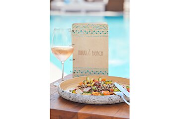 Rose wine, a fresh quinoa salad, and a Nikki Beach menu rest on a wooden table beside a bright blue pool, perfect for a chic resort lunch.