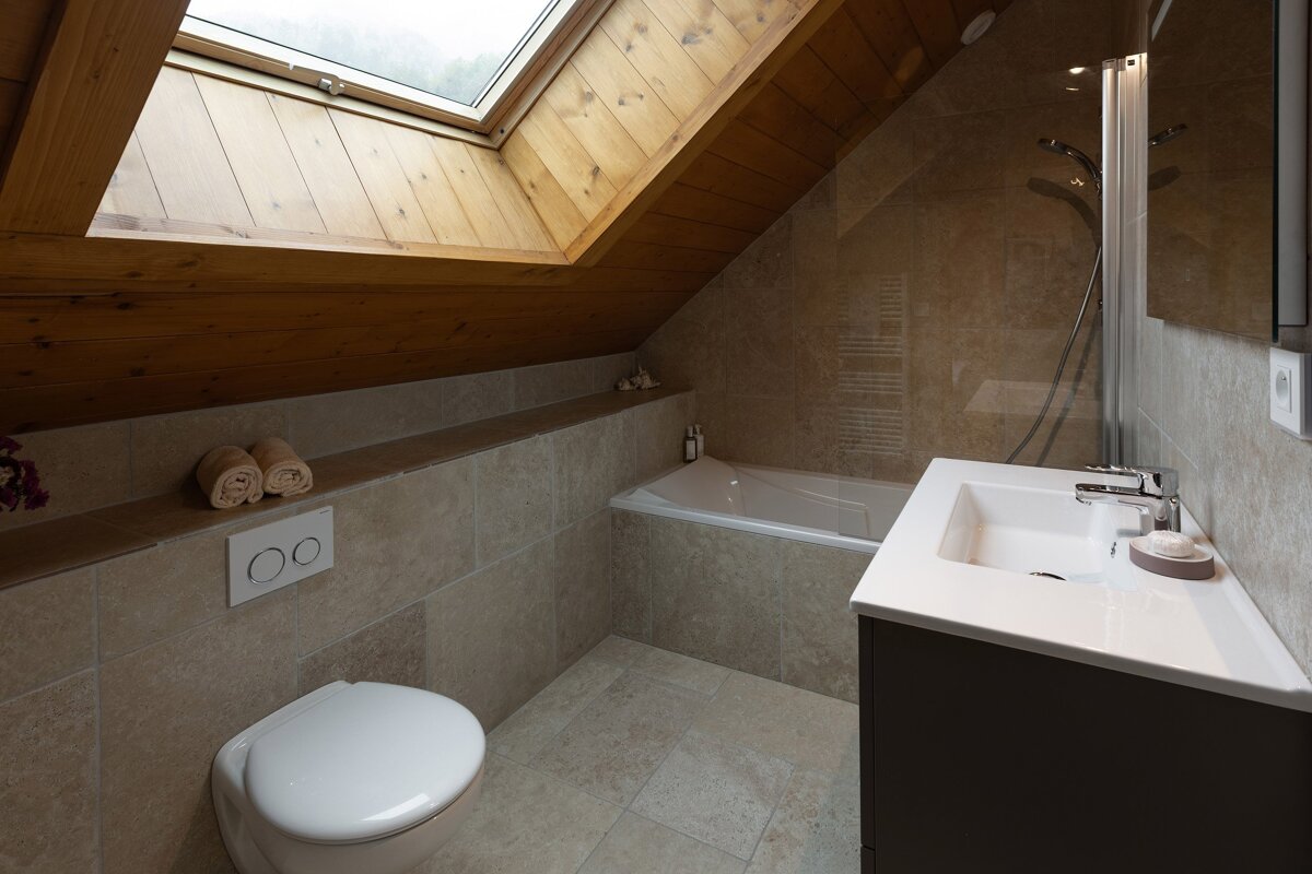 A bright, modern attic bathroom with a skylight, wooden ceiling, beige tiles, bathtub, toilet, and sleek vanity.