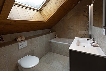 A bright, modern attic bathroom with a skylight, wooden ceiling, beige tiles, bathtub, toilet, and sleek vanity.