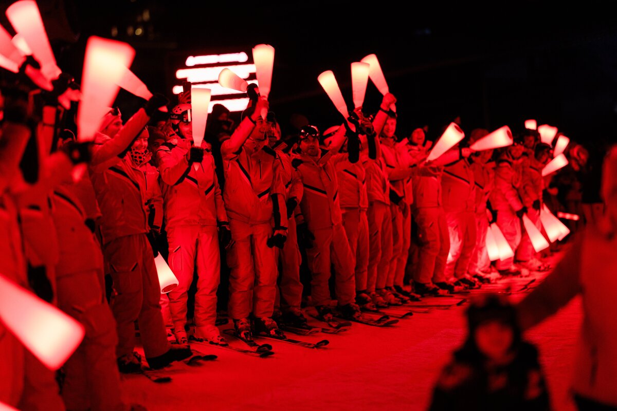 Red-lit skiers in uniform stand in a line at night, holding glowing white, cone-shaped objects high, creating a dramatic, vibrant display.