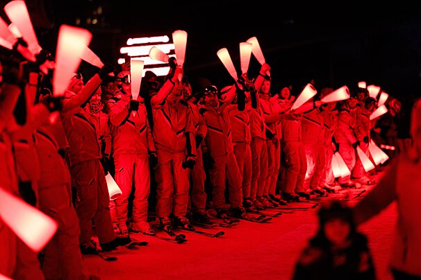 Red-lit skiers in uniform stand in a line at night, holding glowing white, cone-shaped objects high, creating a dramatic, vibrant display.