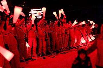 A line of uniformed skiers stands at night, holding glowing white lights aloft, enveloped in an intense red light.