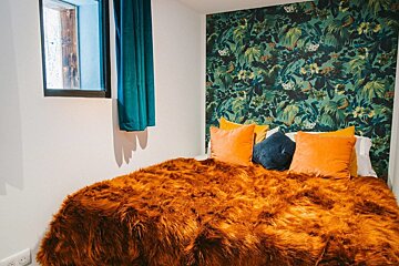 Cozy bedroom featuring a vibrant orange faux fur blanket, tropical leaf wallpaper, and colorful pillows. A window with a teal curtain is visible.