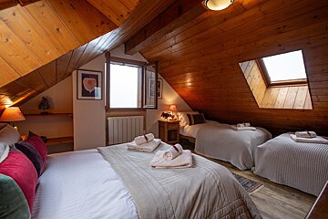 A rustic attic bedroom features warm wood paneling, multiple beds with fresh towels, a dormer window, and a skylight, creating a cozy retreat.