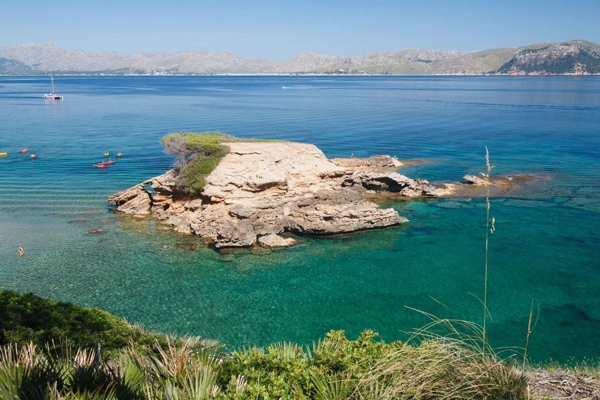 Clear blue-green sea with a rocky island, kayakers, swimmers, and a distant catamaran, backed by mountains under a sunny sky.