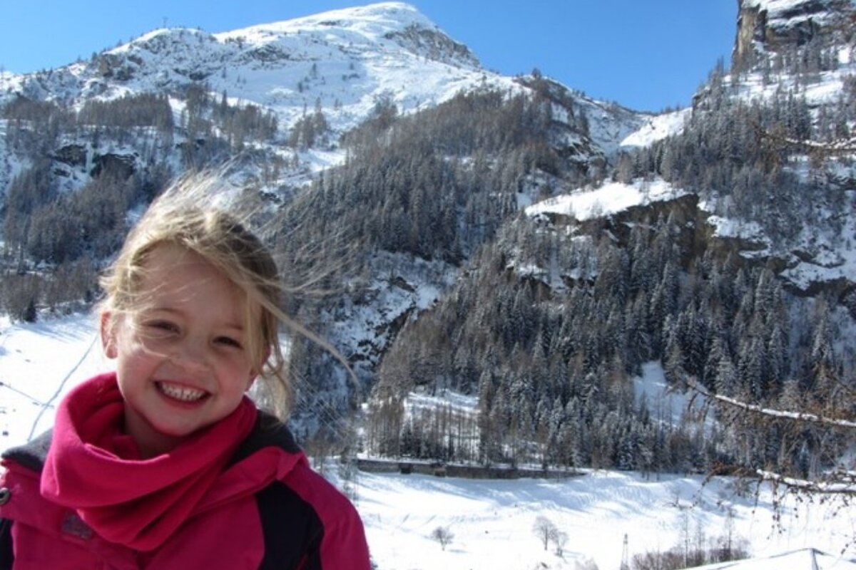 A little girl in a pink jacket smiles in front of a snowy mountain