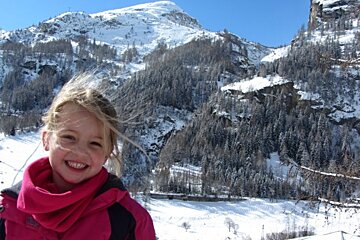 A little girl in a pink jacket smiles in front of a snowy mountain