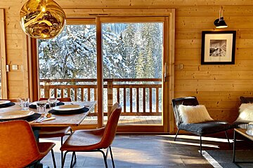 A warm, wood-paneled chalet interior with a dining set and large windows overlooking a snowy forest. Sunlight illuminates the cozy, inviting space.