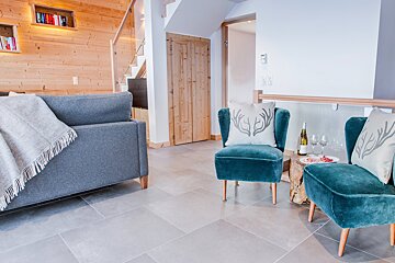 A modern chalet living space with a gray sofa, two teal velvet chairs featuring antler pillows, and a side table with wine and snacks. Light wood walls and tile floors add to the cozy feel.