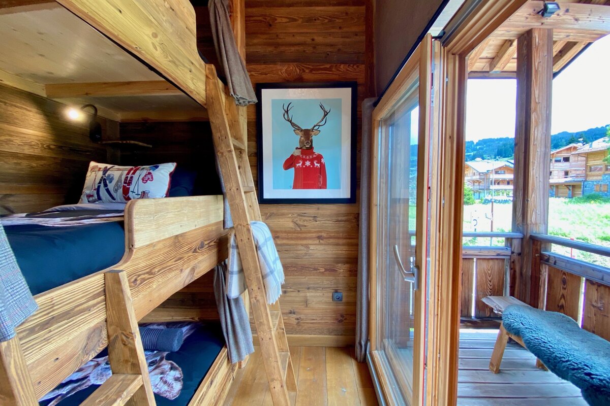A cozy, rustic wooden room features bunk beds, a deer portrait, and an open glass door leading to a balcony with scenic village views.