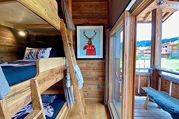 A cozy, rustic wooden room features bunk beds, a deer portrait, and an open glass door leading to a balcony with scenic village views.