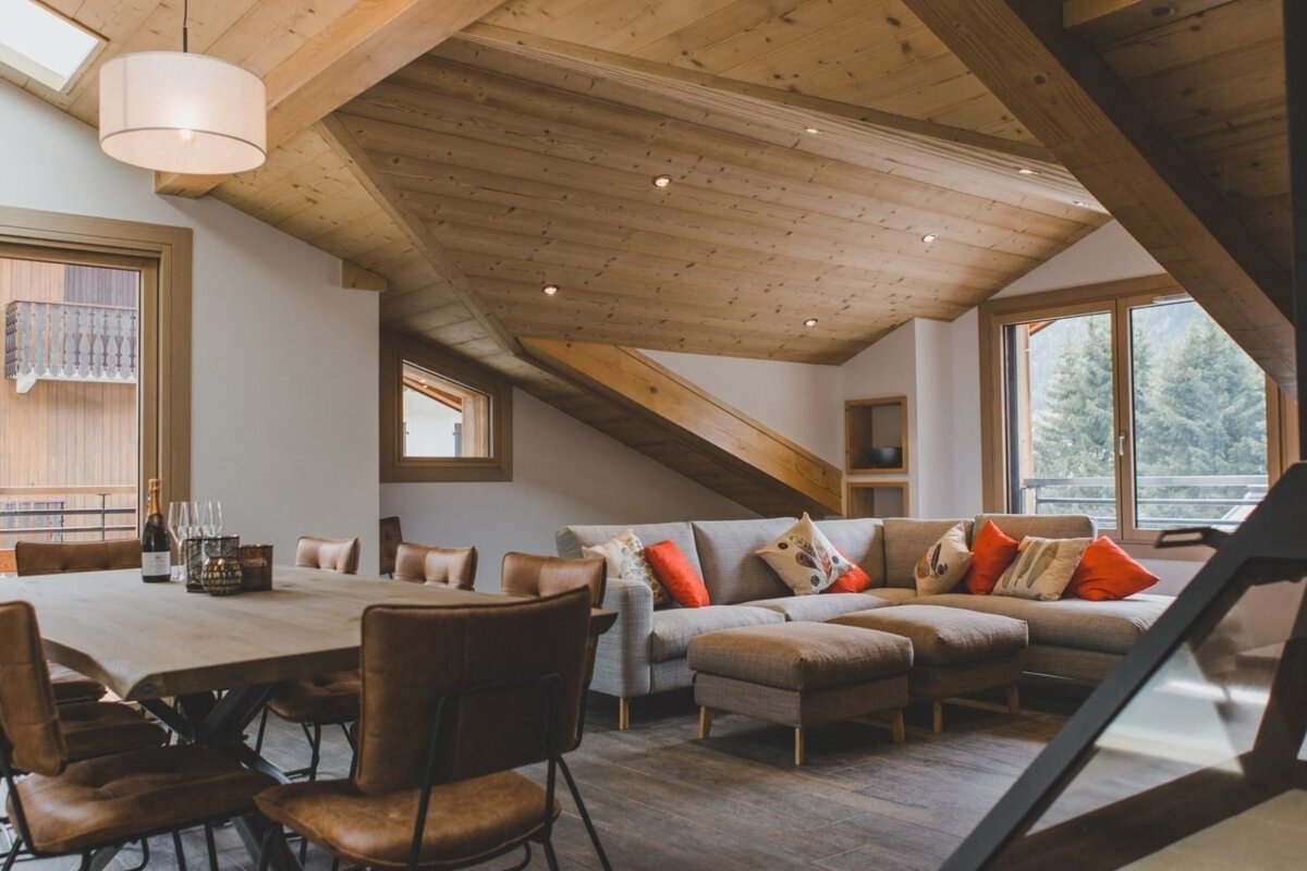 A cozy, modern chalet-style living and dining room. It features a vaulted wooden ceiling, exposed beams, a large sectional sofa, and a rustic dining table, with windows offering forest views.