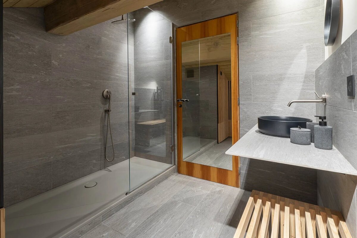 Stylish modern bathroom with grey tiled walls/floor, a glass shower enclosure, a wooden door, and a sleek black sink.