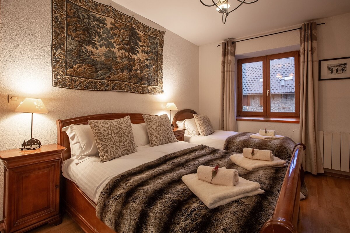 A warm, inviting bedroom with a large bed and a single bed, featuring a scenic tapestry, wooden furniture, and soft lighting for a cozy feel.