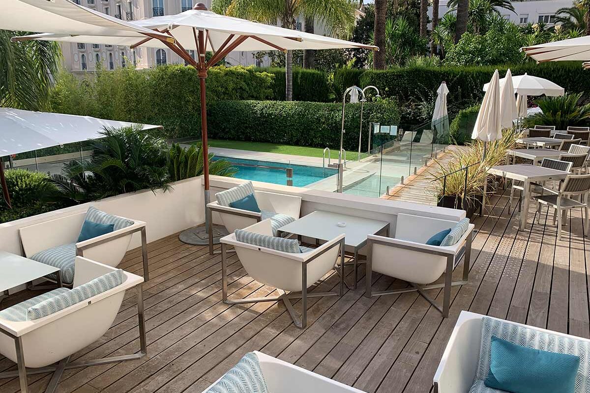 A deck with chairs tables and umbrellas with a pool in the background