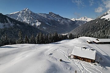 Panoramic winter scene of snow-covered mountains, pine forests, and traditional alpine cabins with ski tracks, under a clear blue sky.