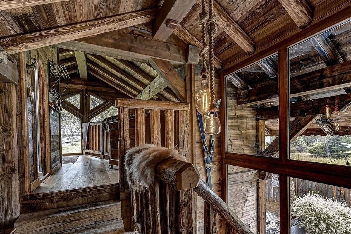 A rustic, multi-level wooden interior features exposed beams, rope lights, skis, and fur on a log railing, with large windows overlooking nature.
