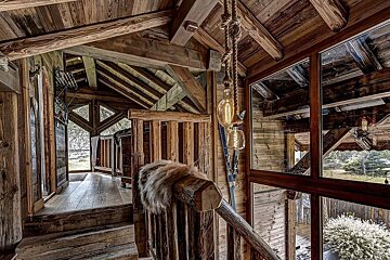 A rustic, multi-level wooden interior features exposed beams, rope lights, skis, and fur on a log railing, with large windows overlooking nature.