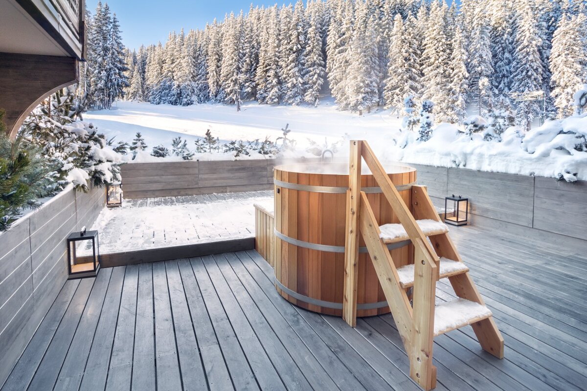A wooden hot tub on a deck with a snowy forest in the background