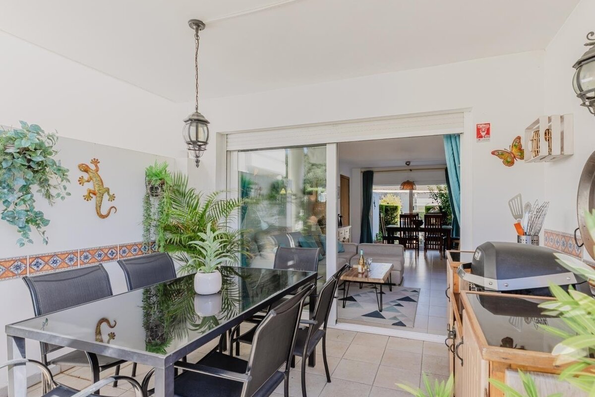 A bright patio with a dining table, BBQ, plants, and lizard decor, seamlessly connecting to an indoor living and dining area through sliding glass doors.