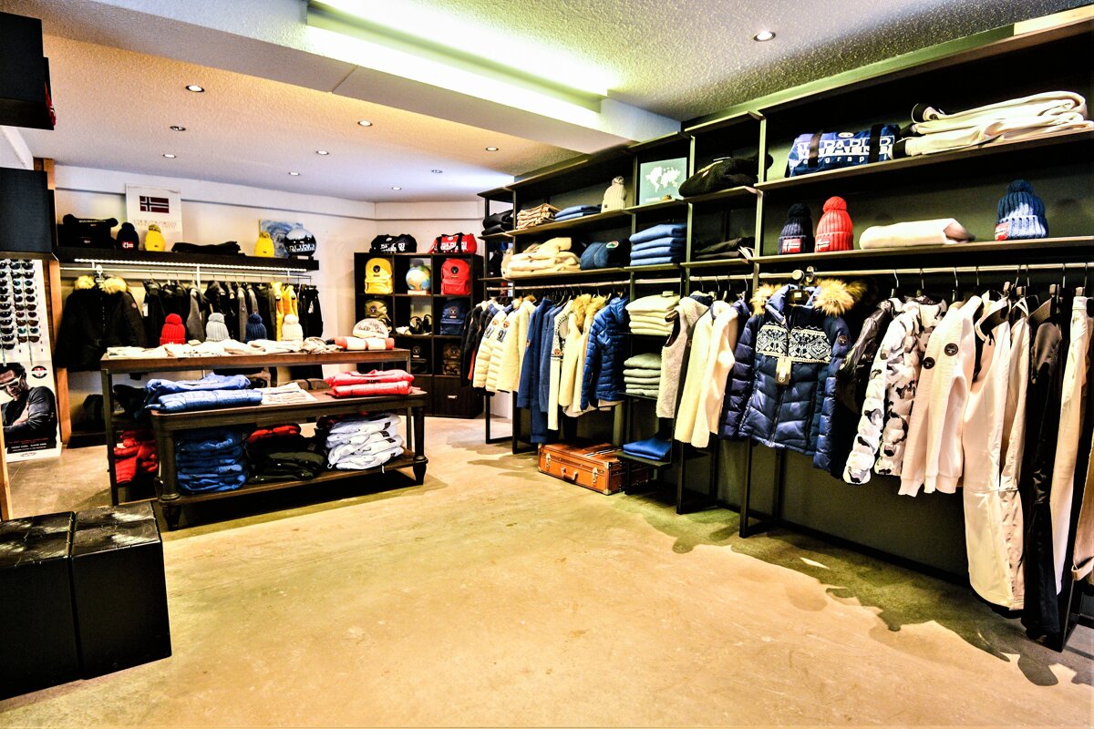 A brightly lit clothing store showcases racks of jackets, folded apparel, hats, and bags on shelves and tables, creating a neat retail display.