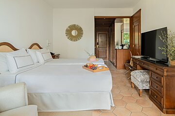 A bright hotel room features twin beds with a breakfast tray, dark wood furniture, a TV, and hexagonal tiled flooring, creating a cozy yet elegant space.