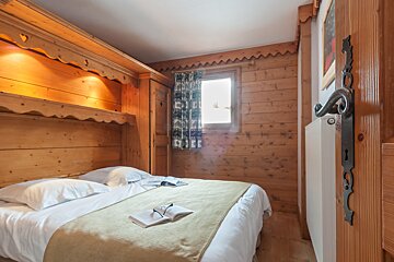 A cozy, rustic wood-paneled bedroom features a bed with white linen, an open book, and a window with patterned curtains. Warm, inviting alpine style.