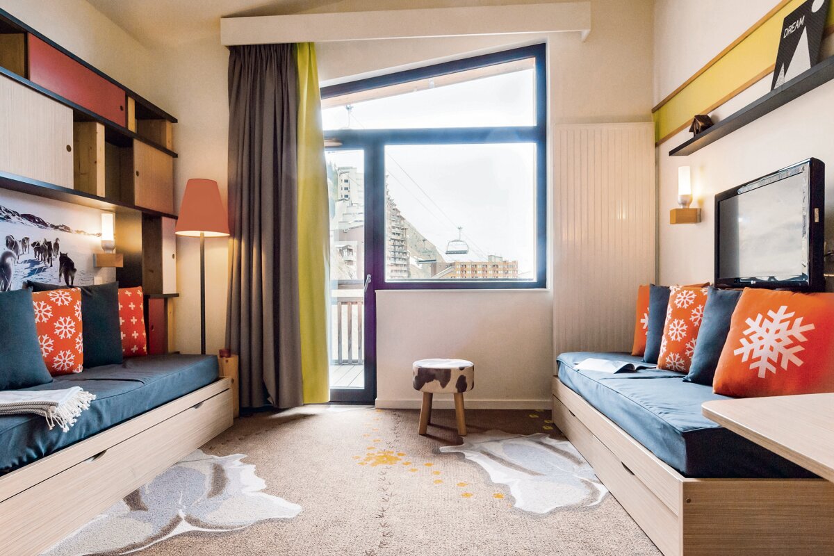 Cozy ski resort room with two daybeds, orange snowflake pillows, and a large window looking out at snowy mountains and a lift.