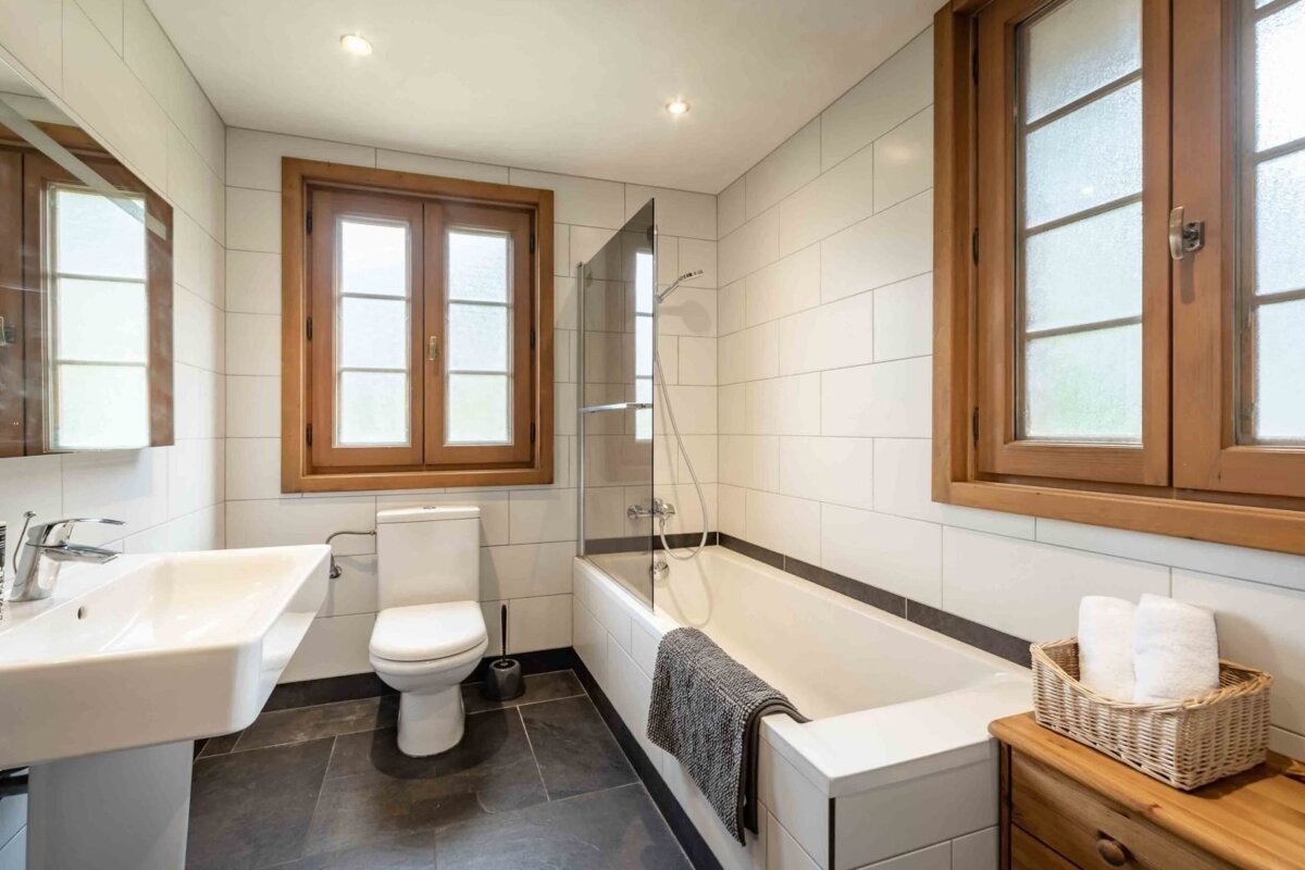 A bright, clean bathroom featuring white tiled walls, a dark floor, and two prominent wooden windows. Includes a bathtub with shower, toilet, and sink.