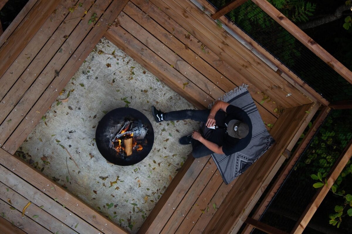 Overhead shot of a person relaxing on a wooden deck next to a fire pit, holding a drink. Scattered leaves are on the ground.