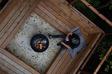 Overhead shot of a person relaxing on a wooden deck next to a fire pit, holding a drink. Scattered leaves are on the ground.