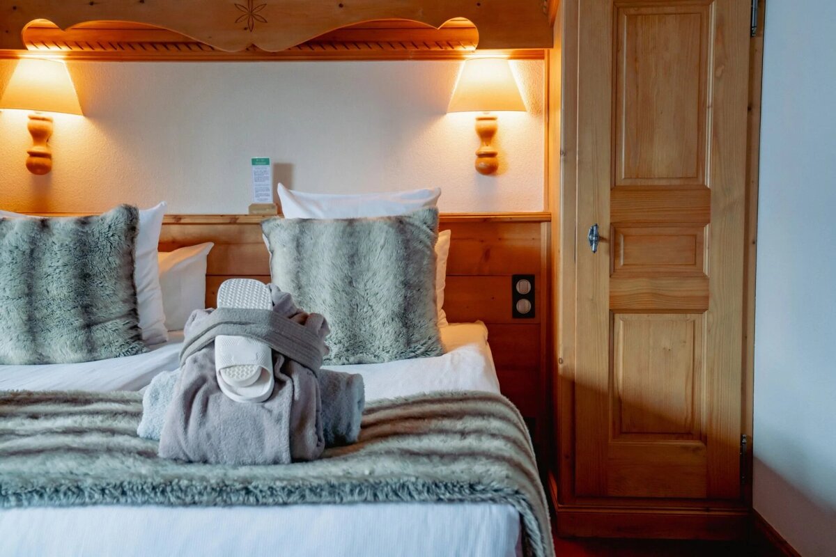 A cozy bedroom featuring a wooden headboard, faux fur pillows and throw, warm lamps, and a robe with slippers neatly placed on the bed.