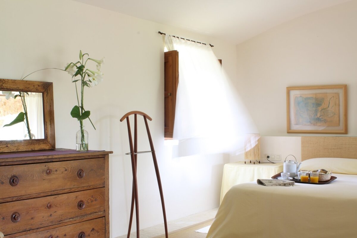 A bright, serene bedroom with white walls, a wooden dresser with flowers, a valet stand, and breakfast on the bed. Sunlight streams through a sheer curtain.