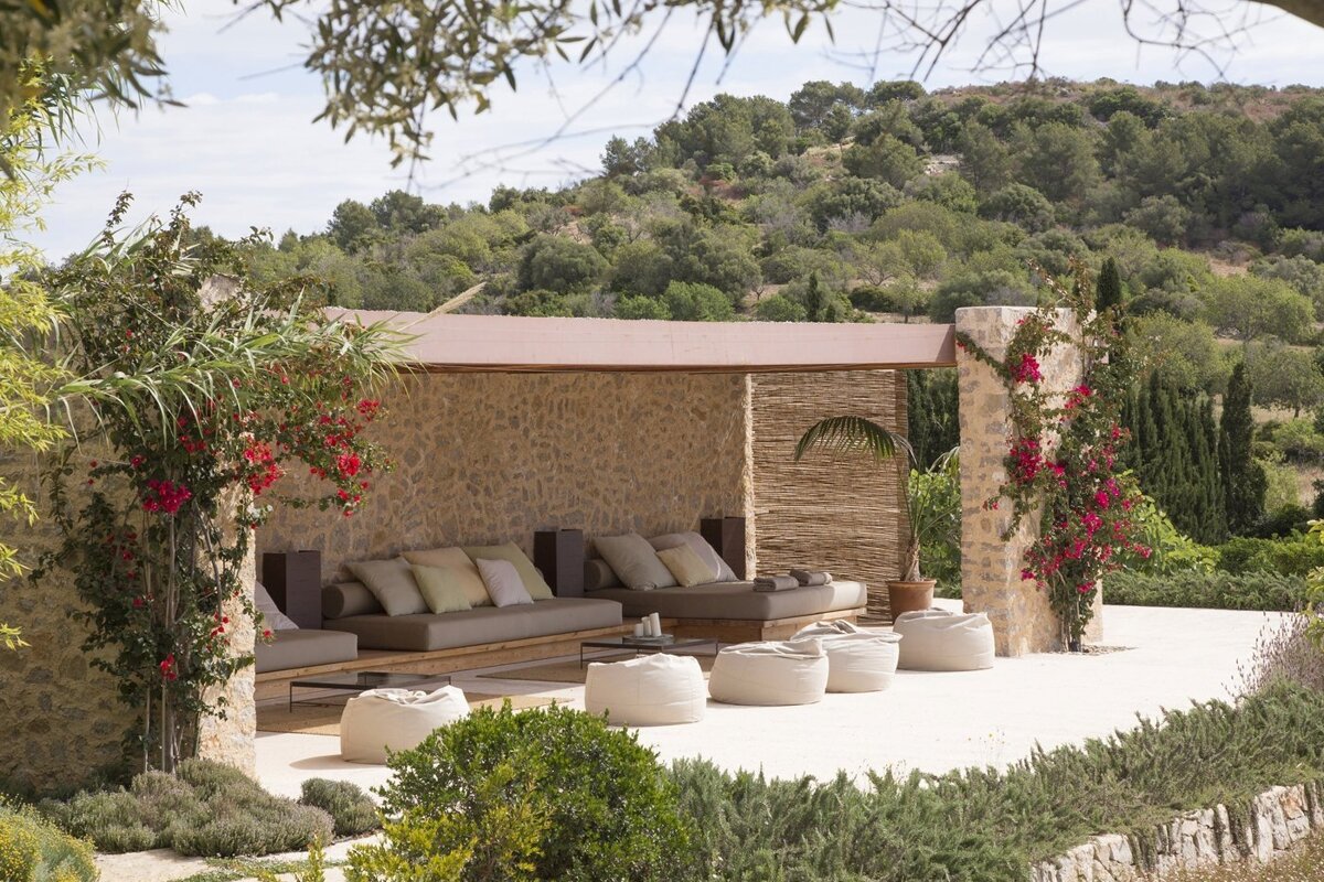 An inviting outdoor lounge with stone walls, cushioned seating, and white poufs, nestled amidst green hills and vibrant bougainvillea.