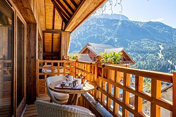 Wooden balcony with breakfast set on a table, overlooking a mountain landscape with chalets.