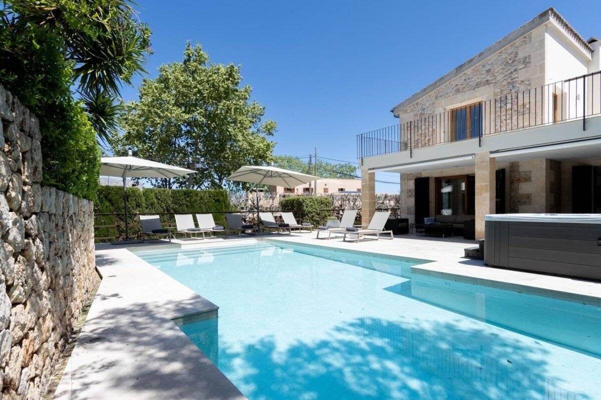 A luxurious stone villa with a clear blue swimming pool, sun loungers, umbrellas, and a jacuzzi under a bright sky.