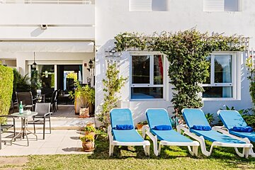 Sunny outdoor area with a white building, patio dining, green lawn, and blue sun loungers. Vines adorn the walls and windows.