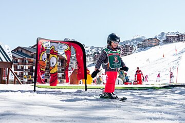 A young child in ski gear, including a helmet and goggles, stands on skis on a sunny mountain slope, near a playful red arch.